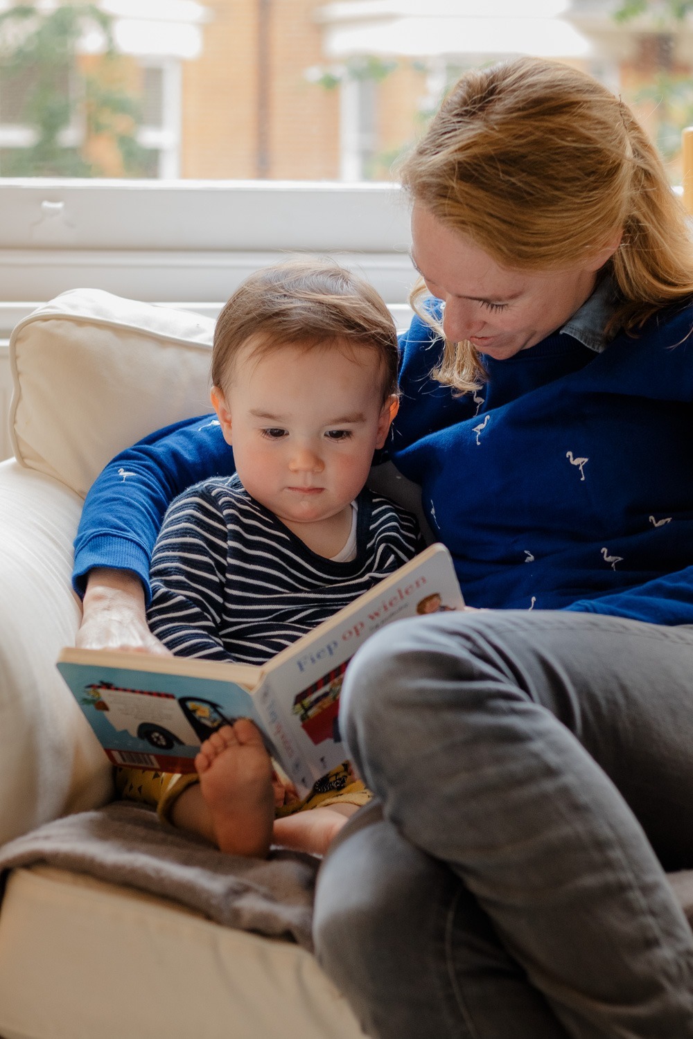 Homestory, kleiner Junge sitzt in großem weißen Sessel, gehalten von der Mutter im blauen Pullover und schaut in ein Buch