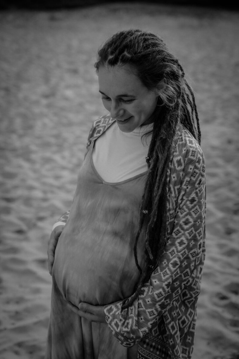 Schwangerschaftsfotografin in Bremen: Unvergessliche Momente festhalten 10 Schwarz-Weiss Porträtaufnahme einer Frau auf Babybauch schauend, mit Dünen im Hintergrund