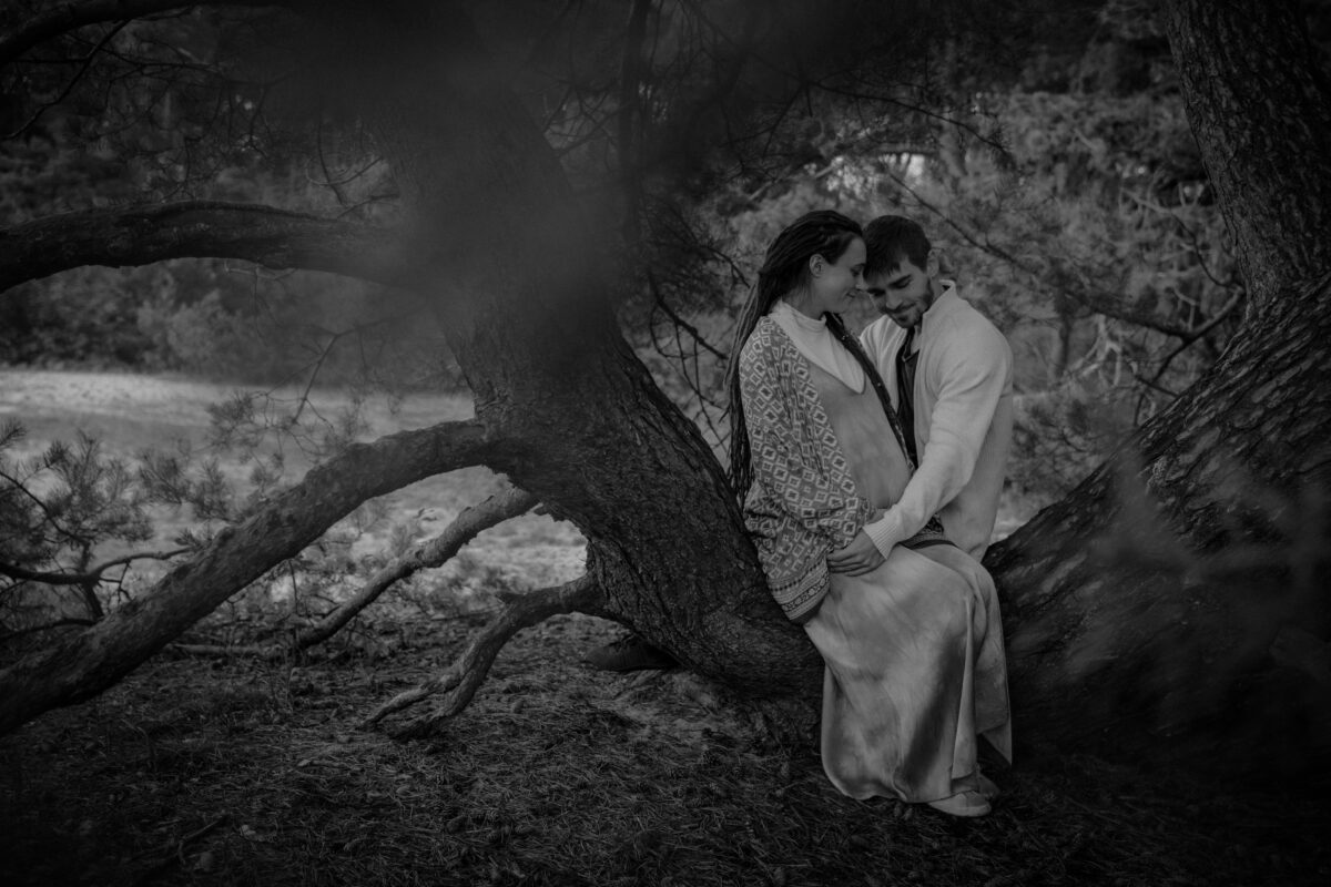 Schwarz-Weiss Aufnahme Paar in den Dünen mit Wald im Hintergrund, Frau mit Babybauch, Mann hält Bauch, Paar sitzt auf Baumstamm, innig verbunden